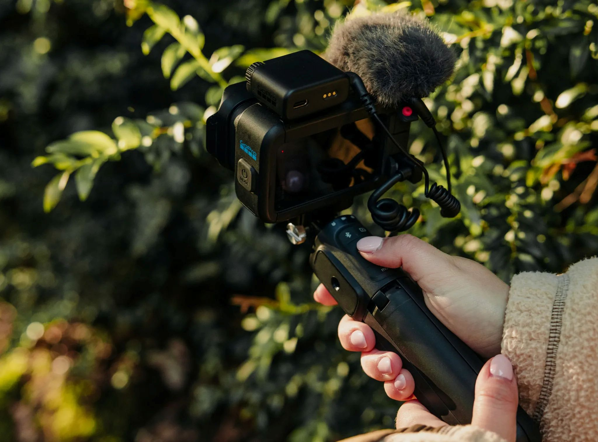 Eine Hand hält eine Actioncam mit Mikrofon auf einem Griff vor grünen Blättern.