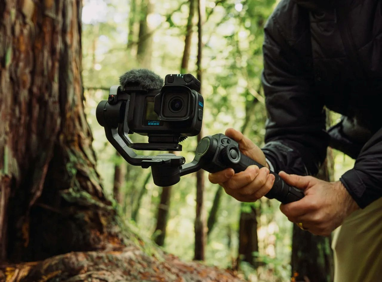 Eine Person hält einen schwarzen Kamera-Gimbal in einem grünen Wald mit hohen Bäumen im Hintergrund.