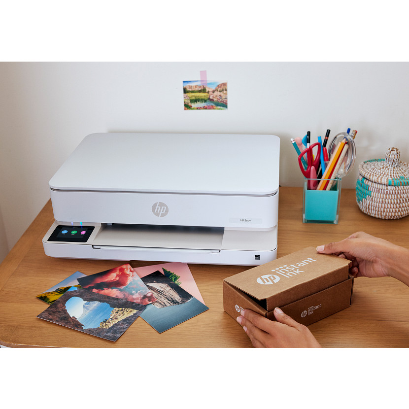 Ein weißer HP-Drucker auf einem Holztisch mit gedruckten Fotos und einer Person, die eine HP Instant Ink-Schachtel öffnet.