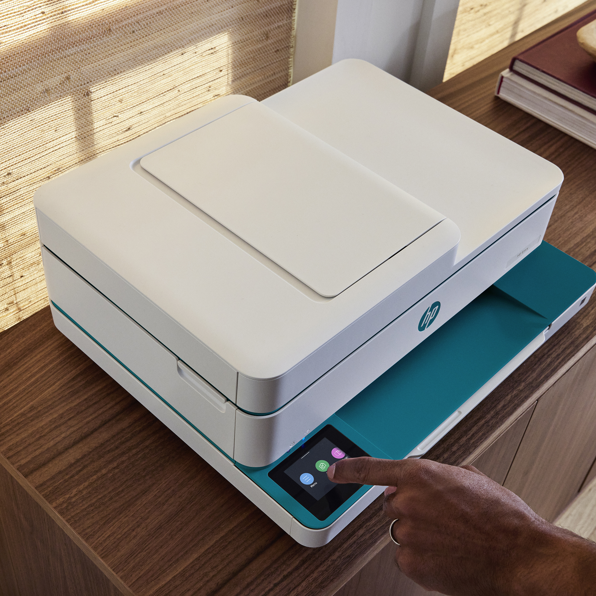 Eine Hand bedient den Touchscreen eines weißen HP Envy Druckers auf einem Holztisch.
