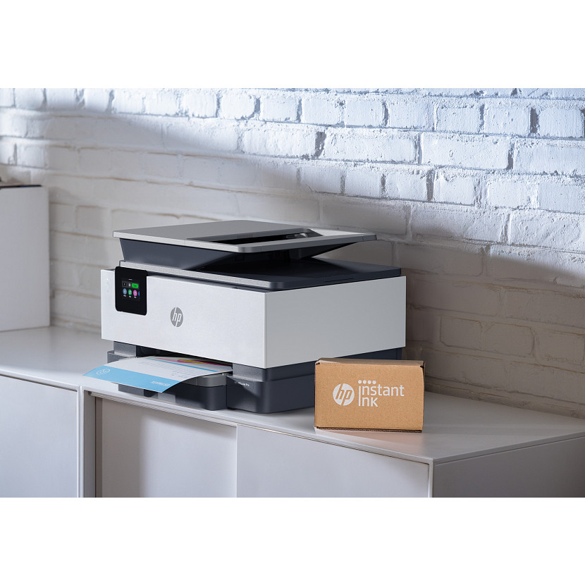 Ein weiß-grauer HP Drucker steht auf einem weißen Schrank vor einer weißen Backsteinwand, daneben ein HP Instant Ink Paket.