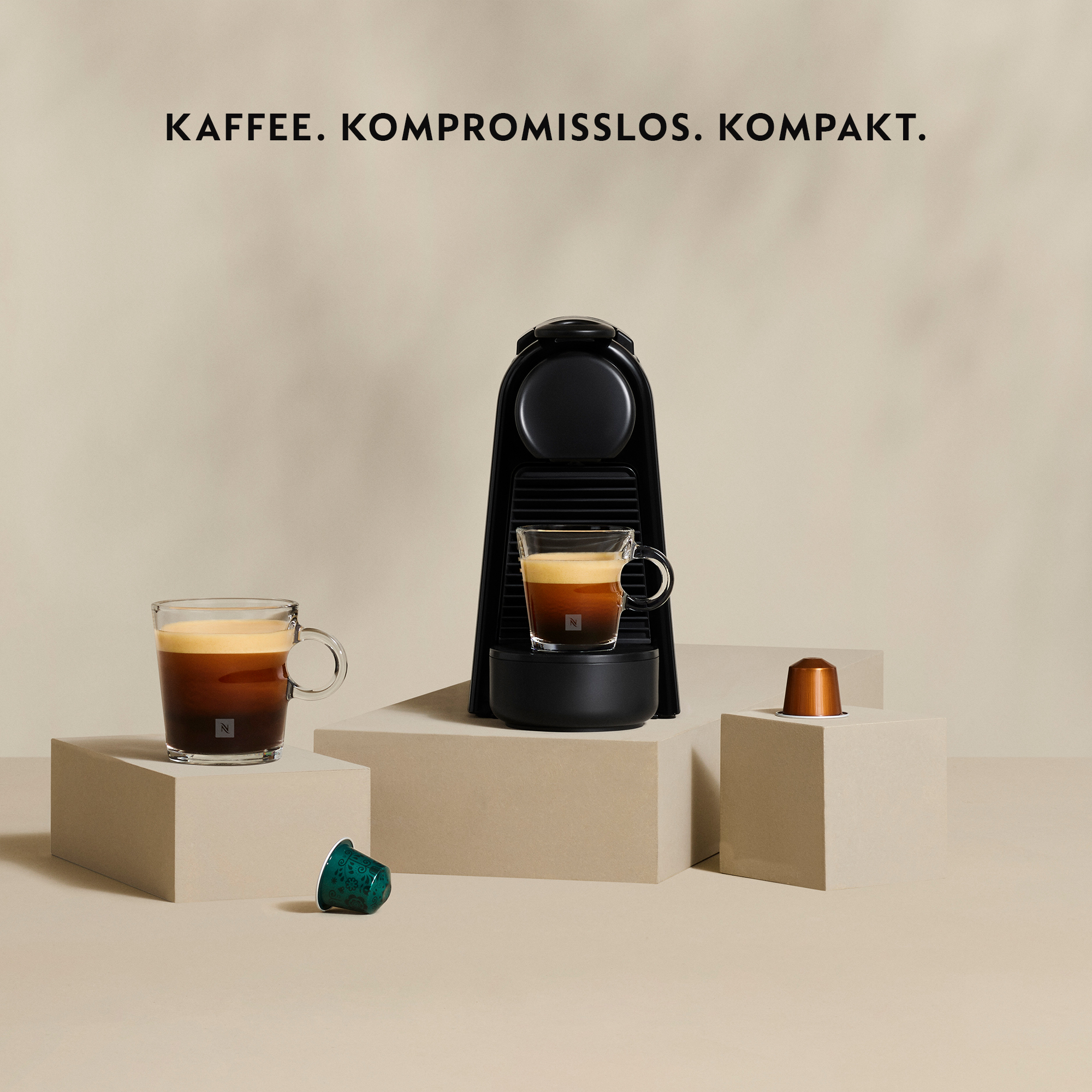Schwarze Kaffeemaschine auf Podesten mit zwei Tassen Kaffee und Kapseln, vor beigem Hintergrund.