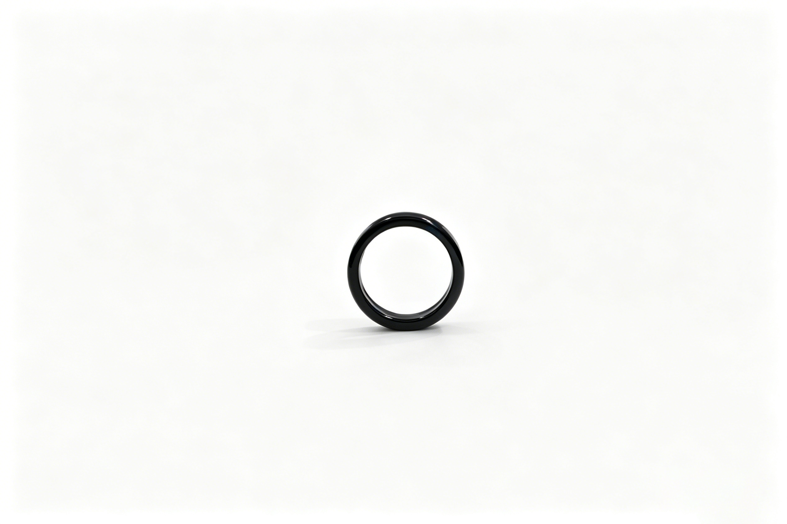 Une bague noire brillante se dresse verticalement sur une surface blanche lumineuse sur un fond neutre.