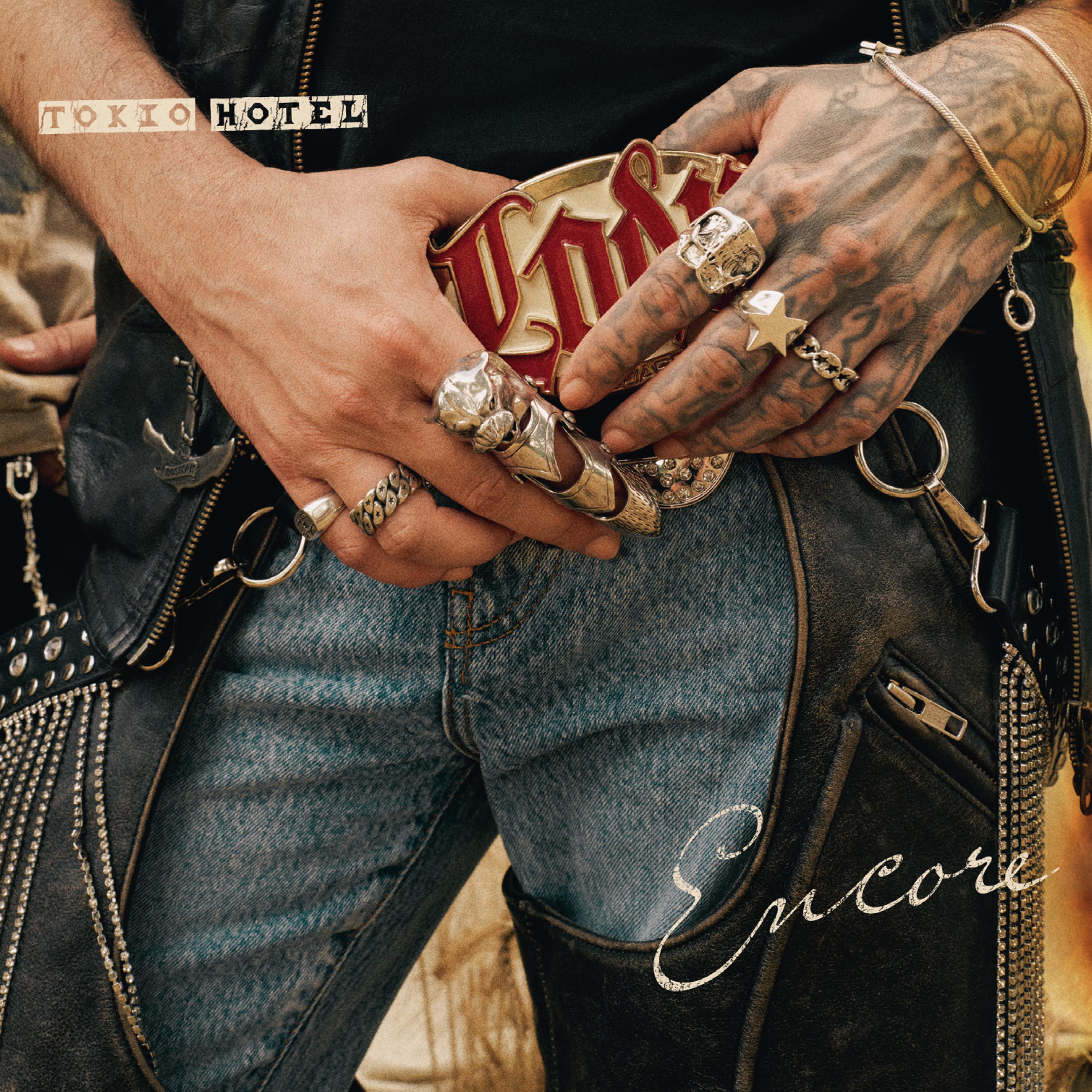 Tätowierte Hände mit Ringen halten eine große Gürtelschnalle vor Jeans und Lederchaps.