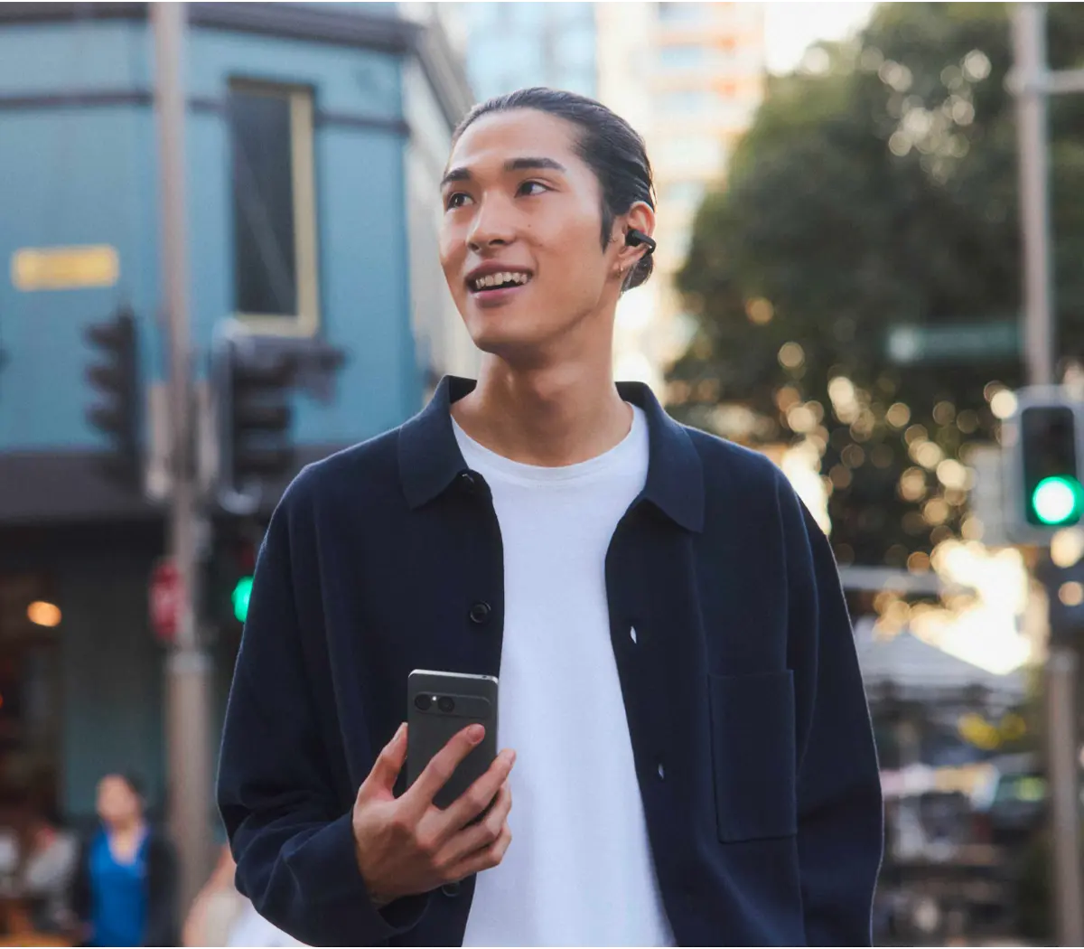 Uzun koyu saçlı, beyaz gömlekli ve koyu ceketli genç bir adam sokakta akıllı telefon tutuyor.