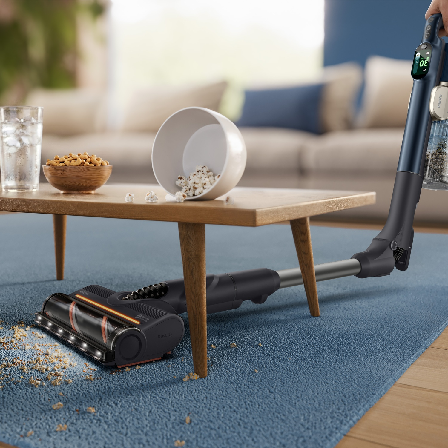 A cordless vacuum cleans carpet crumbs under a wooden coffee table in a living room.
