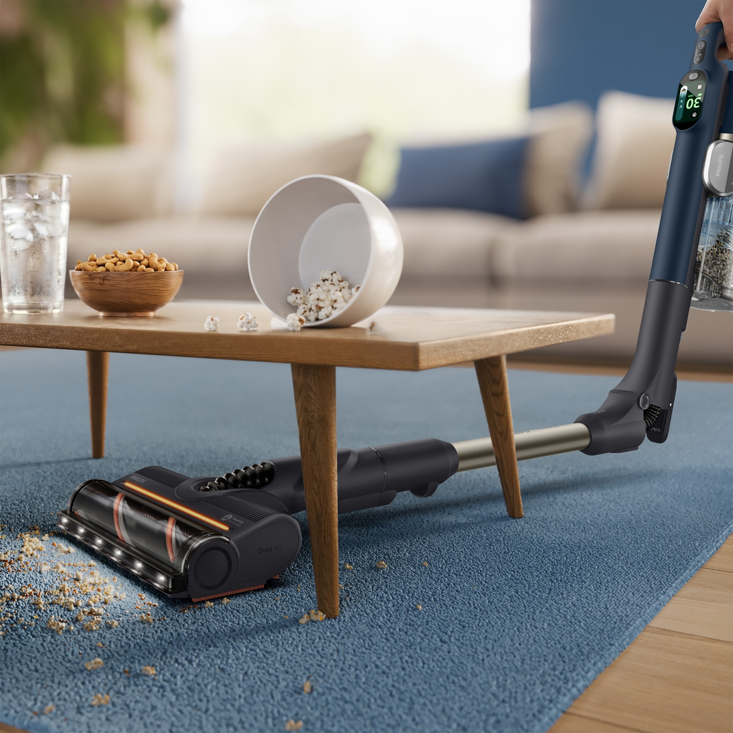 A blue cordless vacuum cleans spilled popcorn and crumbs from a blue rug under a wooden table.