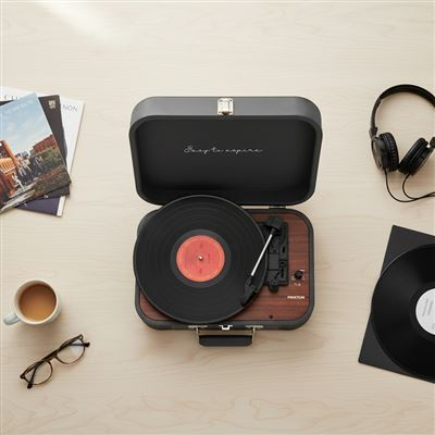 Un tocadiscos de maleta gris en una mesa de madera, rodeado de auriculares, discos de vinilo, gafas y una taza de café.