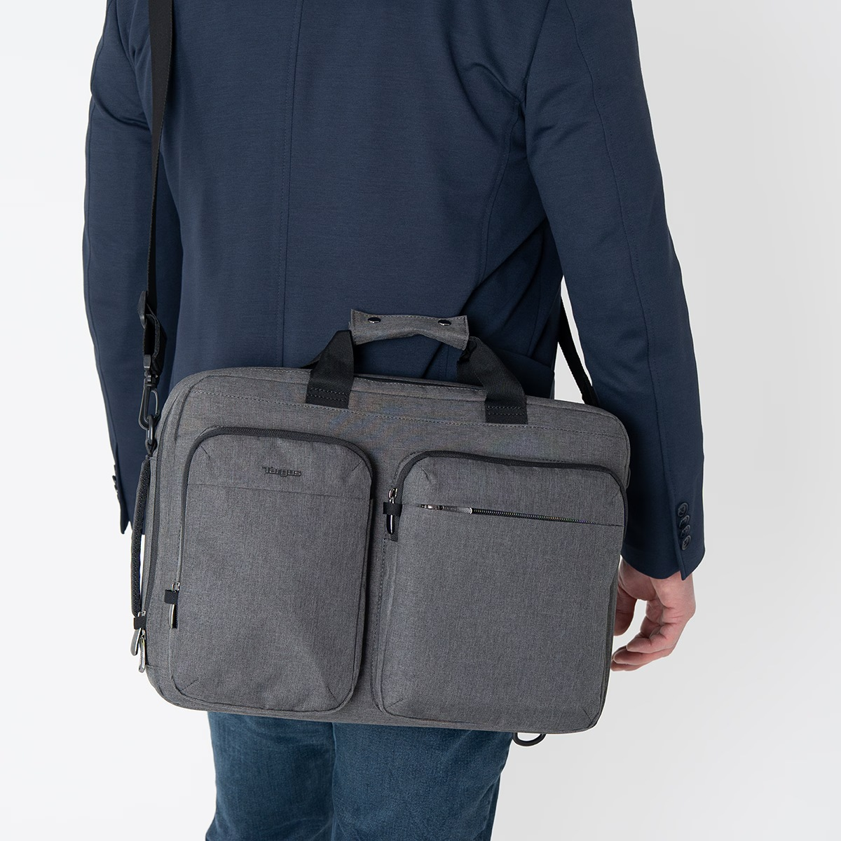 Una persona con chaqueta azul y vaqueros lleva una bolsa gris para portátil con correa.