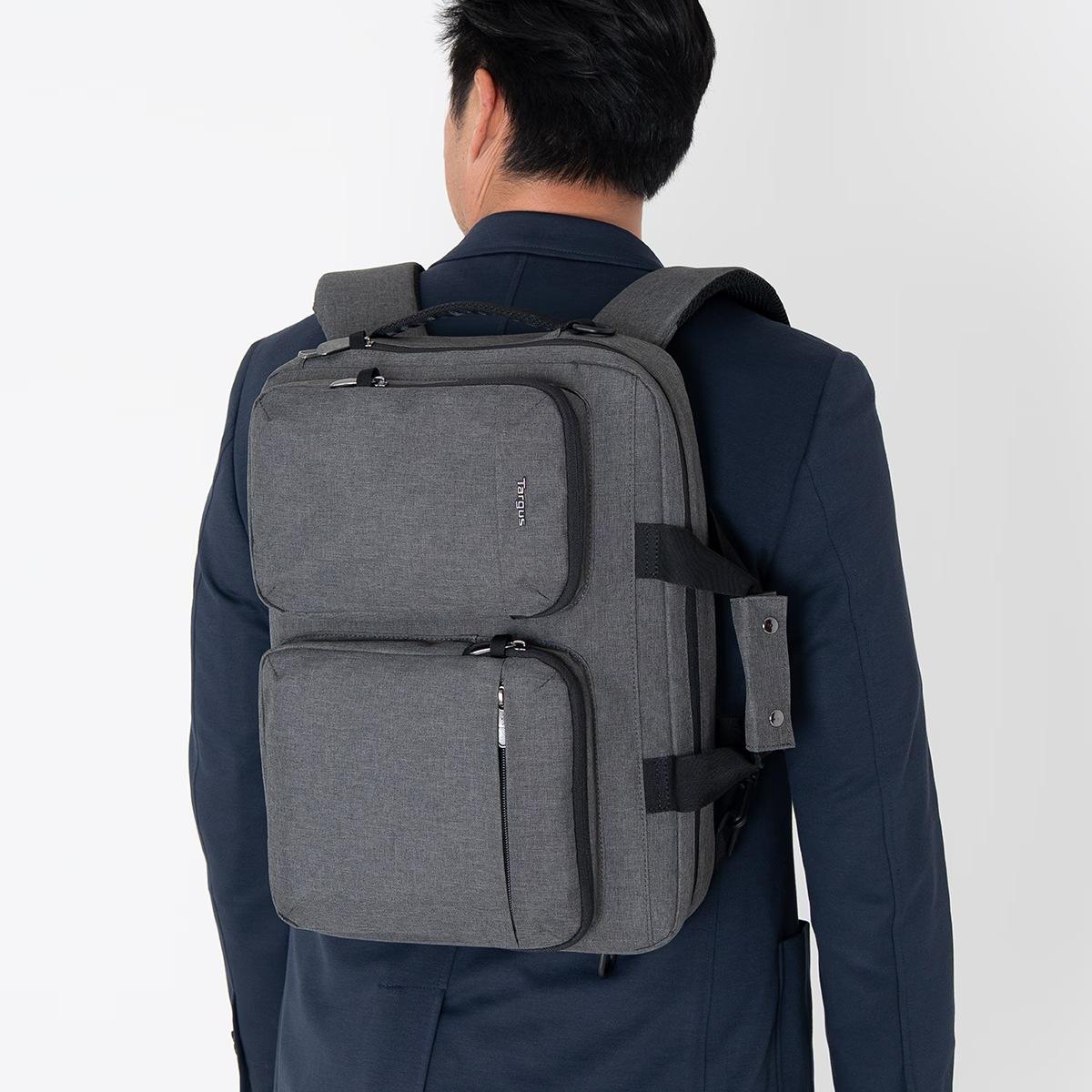 Un hombre con traje azul oscuro lleva una mochila gris Targus con bolsillos frontales en la espalda.