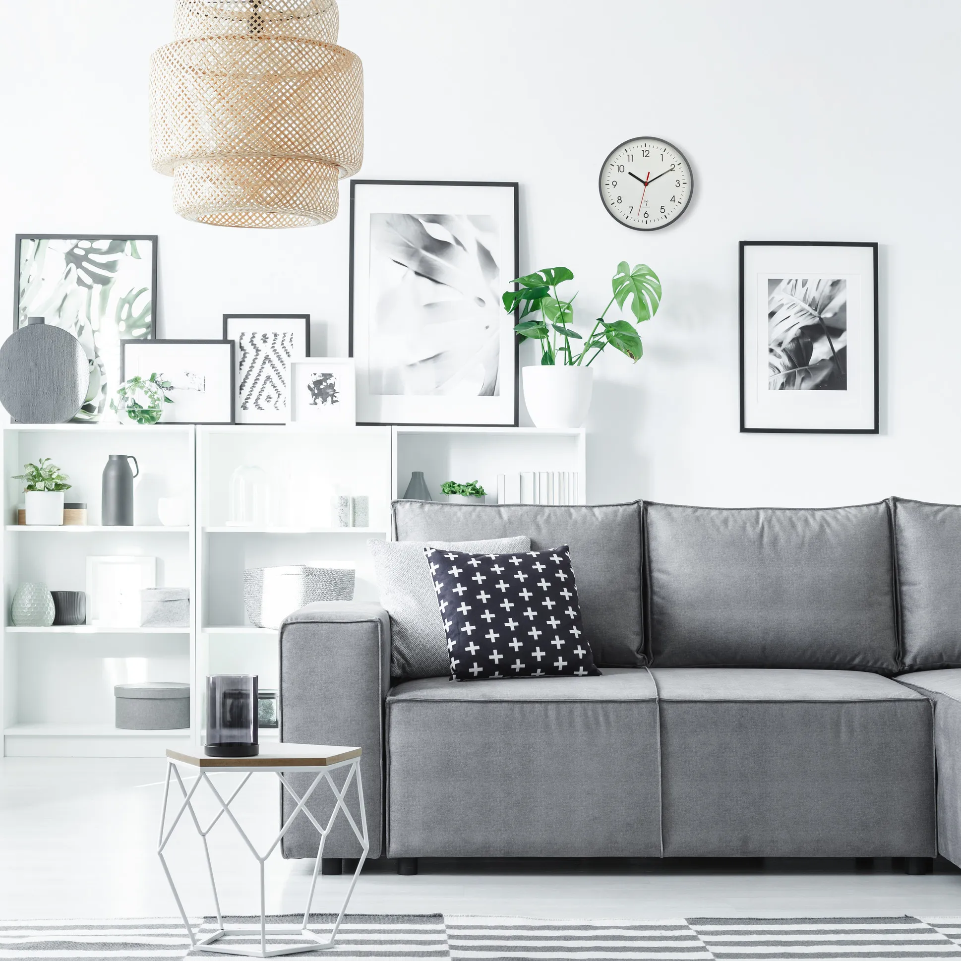 Wohnzimmer mit grauem Sofa, Kissen, Wanduhr, gerahmten Bildern, weißem Regal und einer großen geflochtenen Hängeleuchte.