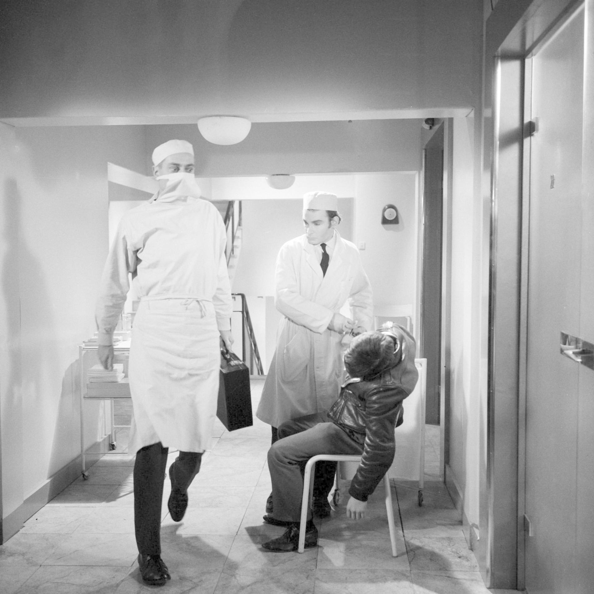 Ein Chirurg in Schutzkleidung geht durch einen Krankenhausflur, während ein Arzt einem Patienten auf einem Stuhl hilft.