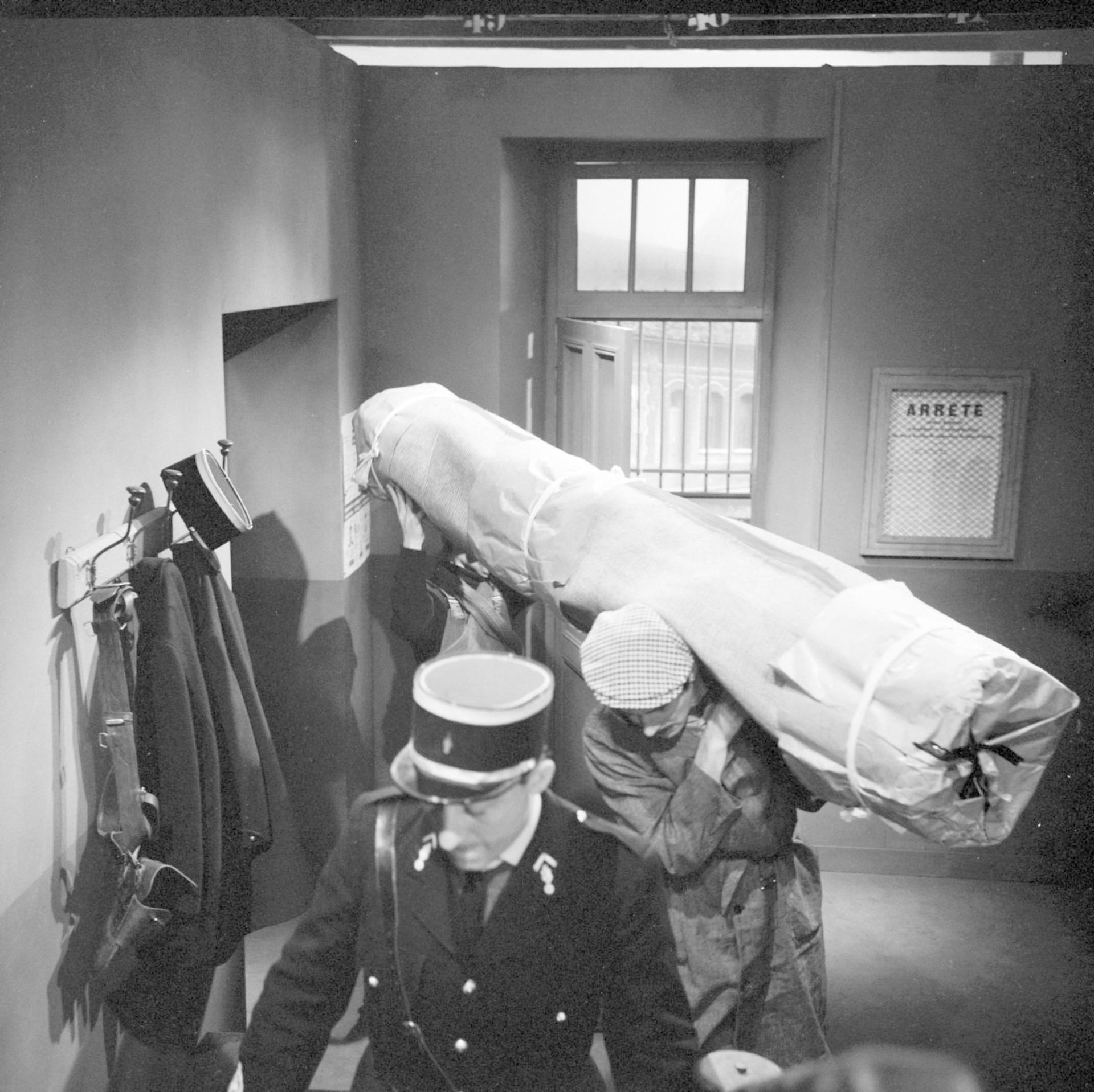 Zwei Männer tragen ein großes, in Papier gewickeltes Paket, gefolgt von einem Polizisten.