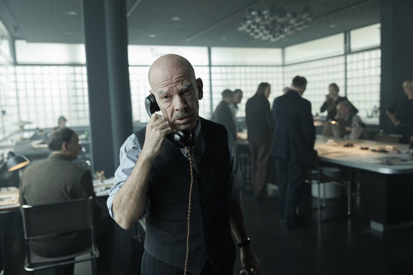Ein kahlköpfiger älterer Mann in Weste telefoniert in einem modernen, hellen Großraumbüro mit Kollegen im Hintergrund.