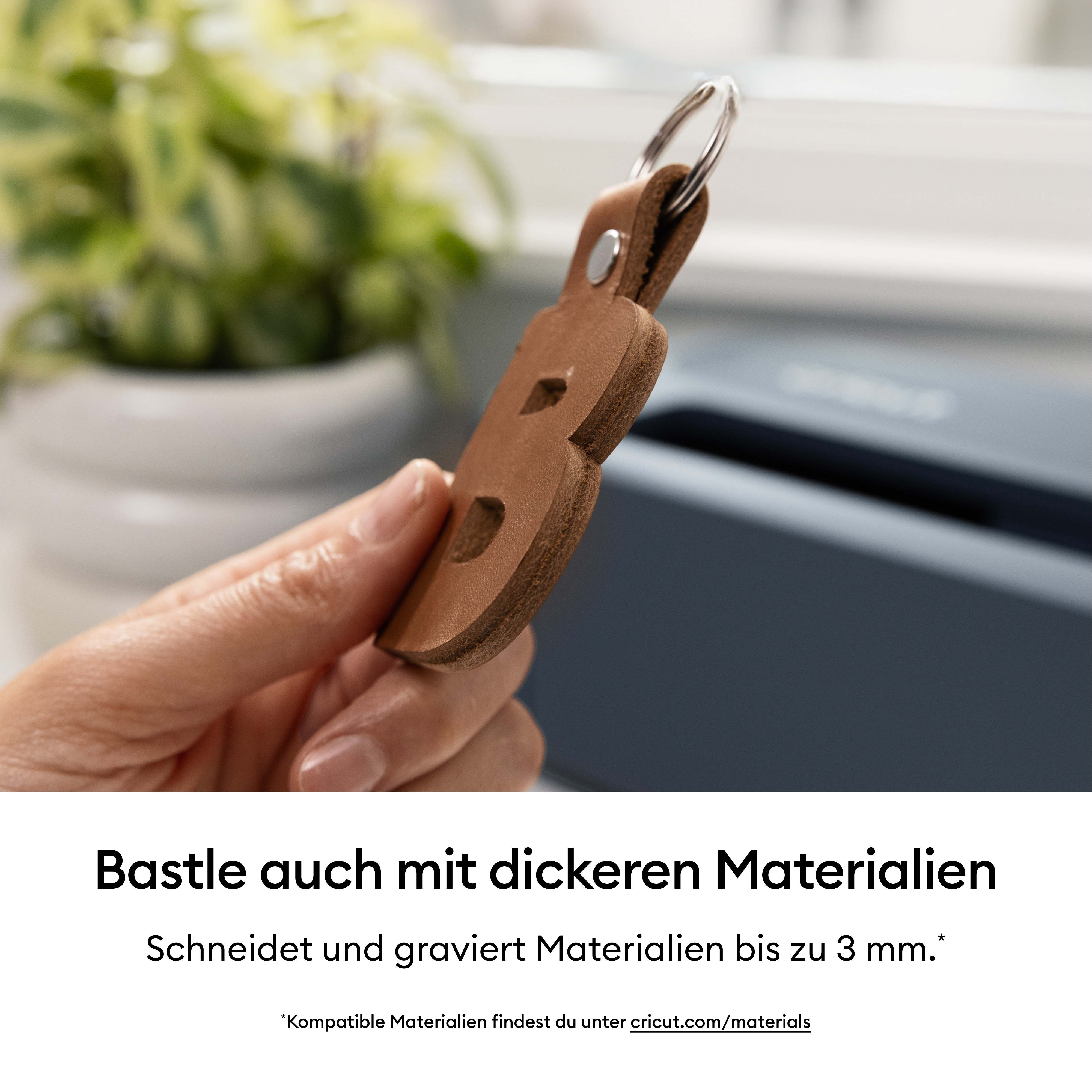 Eine Hand hält einen braunen Lederanhänger mit einem Buchstaben, im Hintergrund eine Pflanze und ein Gerät.