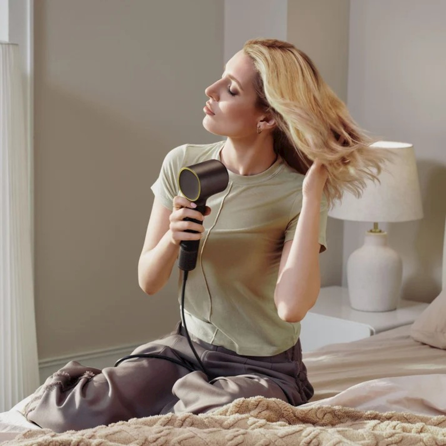 Una donna bionda siede sul letto asciugandosi i capelli con un phon moderno.
