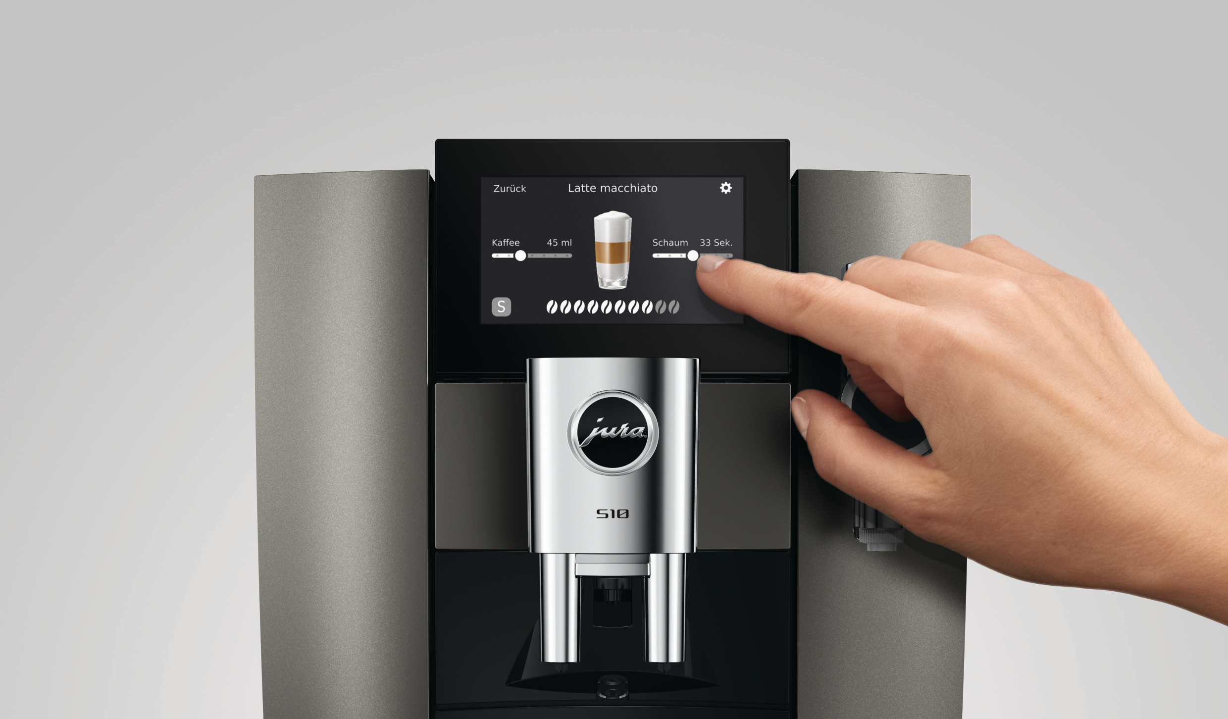 Hand bedient den Touchscreen einer grauen Jura S10 Kaffeemaschine mit Latte-Macchiato-Display.