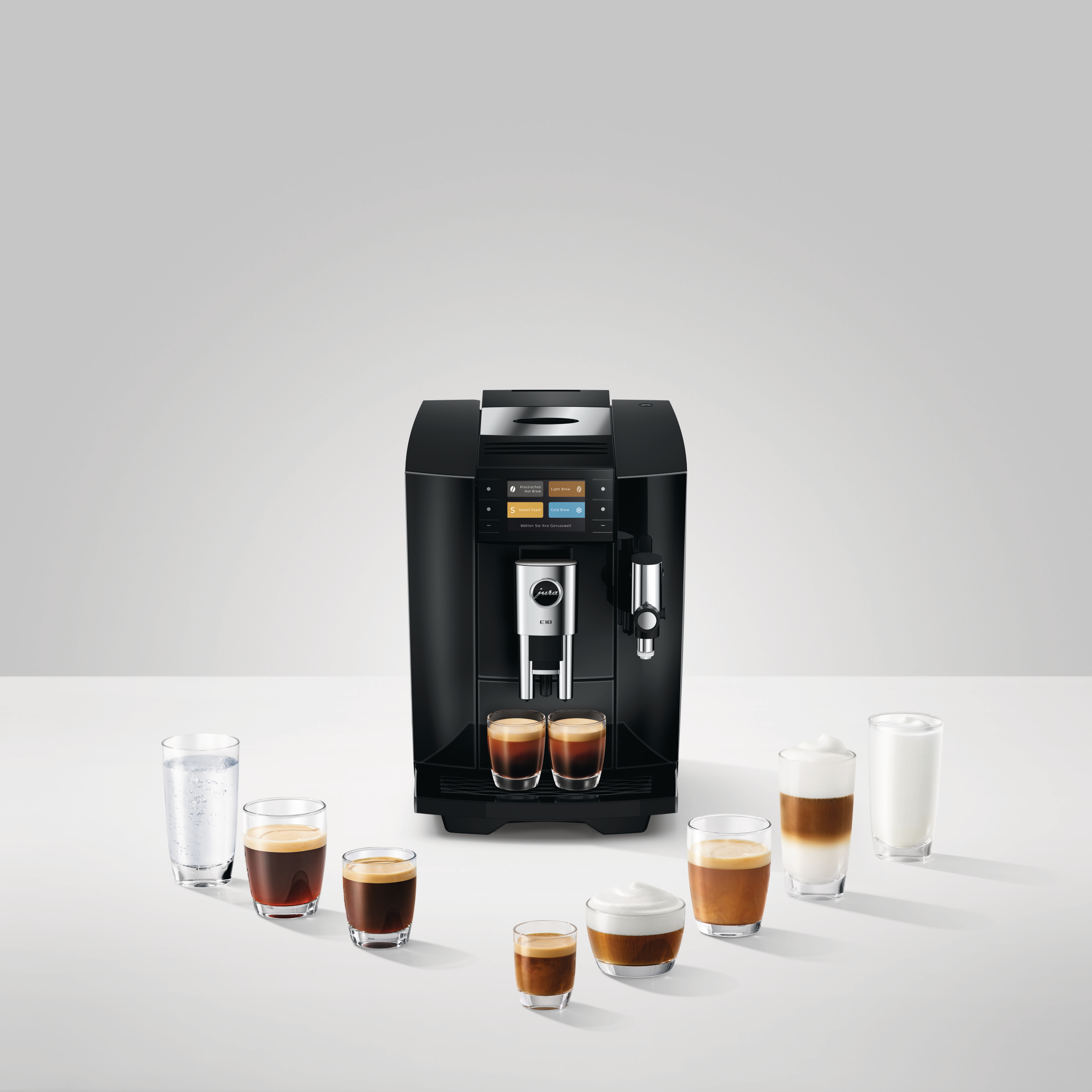 Eine schwarze Jura E8 Kaffeemaschine steht auf weißem Tisch, umgeben von Gläsern mit Kaffee, Milch und Wasser.
