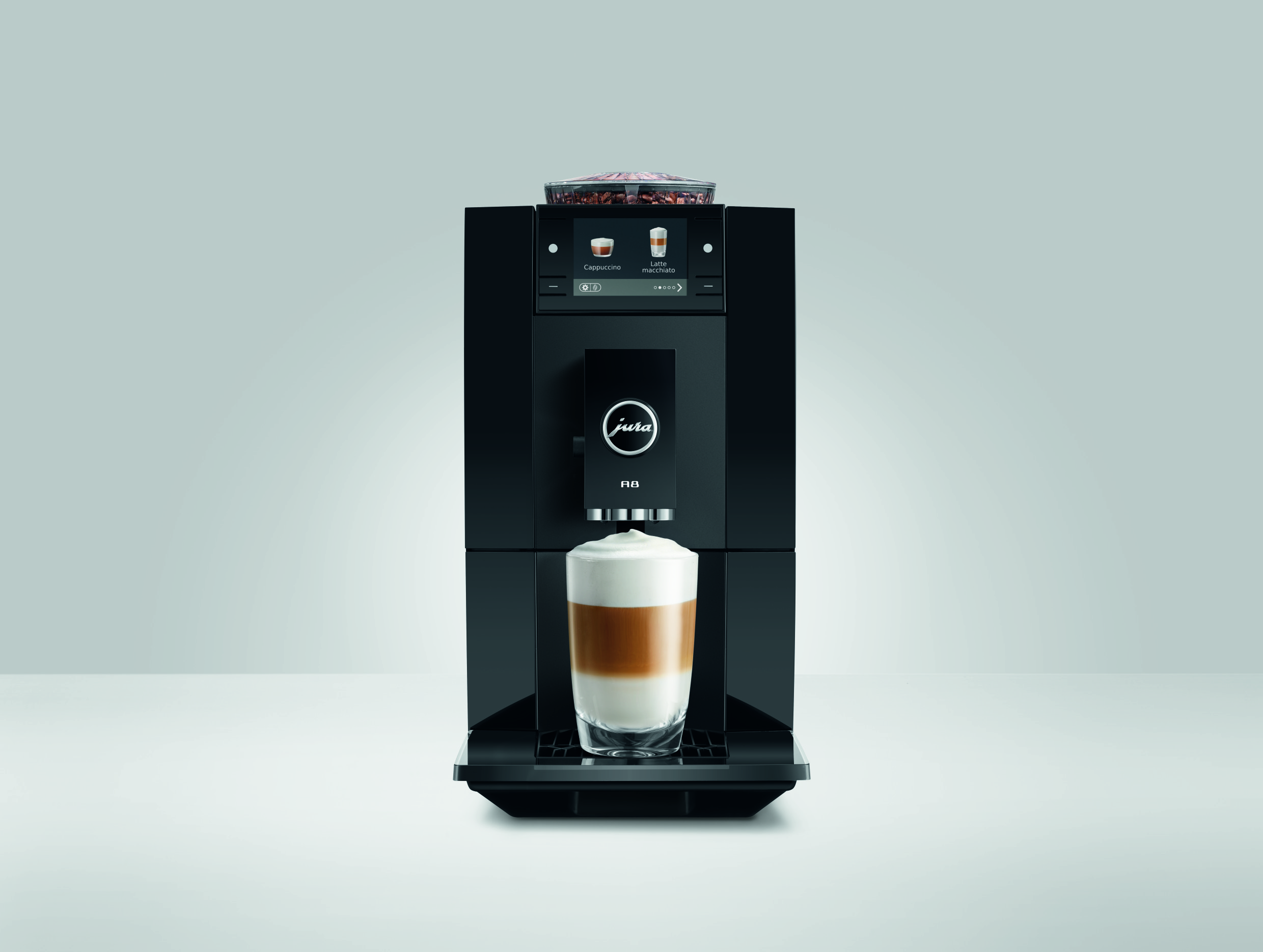 Eine schwarze Jura A8 Kaffeemaschine mit einem frisch zubereiteten Latte Macchiato in einem Glas auf weißem Tisch.