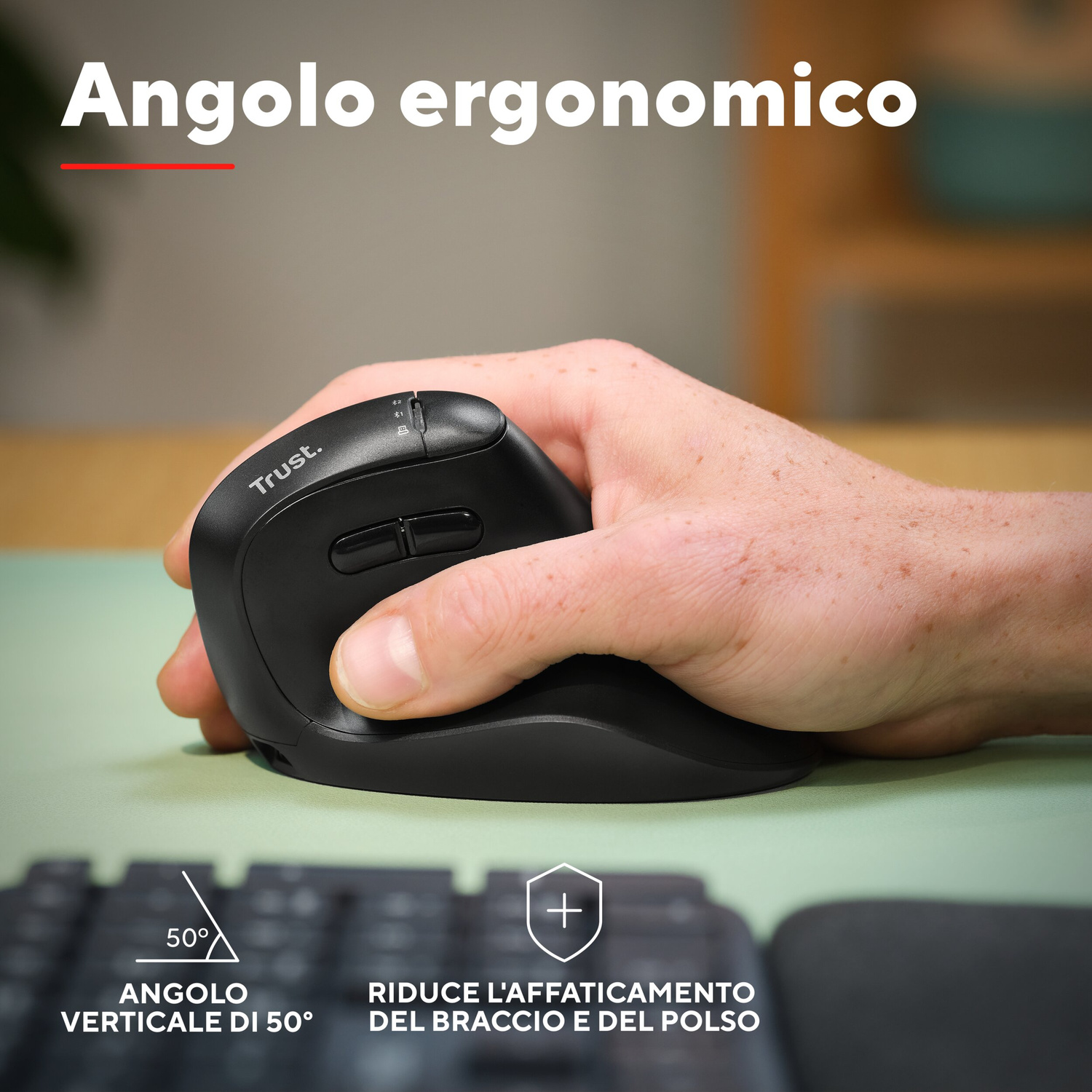 Mano che impugna un mouse verticale Trust nero su tappetino verde con tastiera in primo piano.