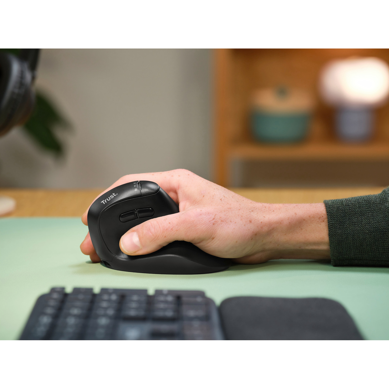 Una mano afferra un mouse per computer ergonomico nero Trust su un tappetino da scrivania verde.