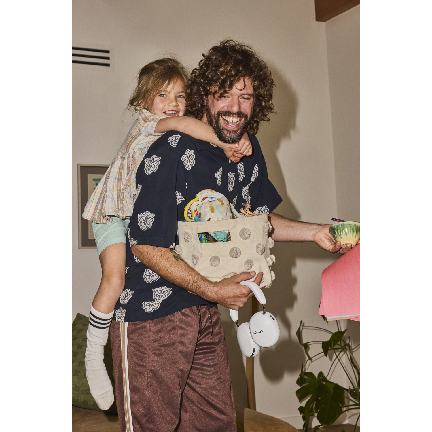 Een gelukkige man draagt een jong meisje op zijn schouders en houdt een tas en koptelefoon vast.