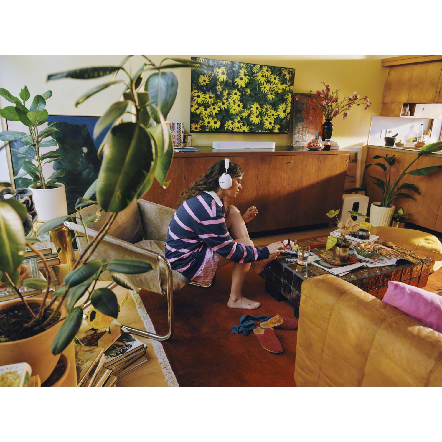 Vrouw met koptelefoon zit in een lichte, plantenrijke woonkamer haar nagels te lakken.