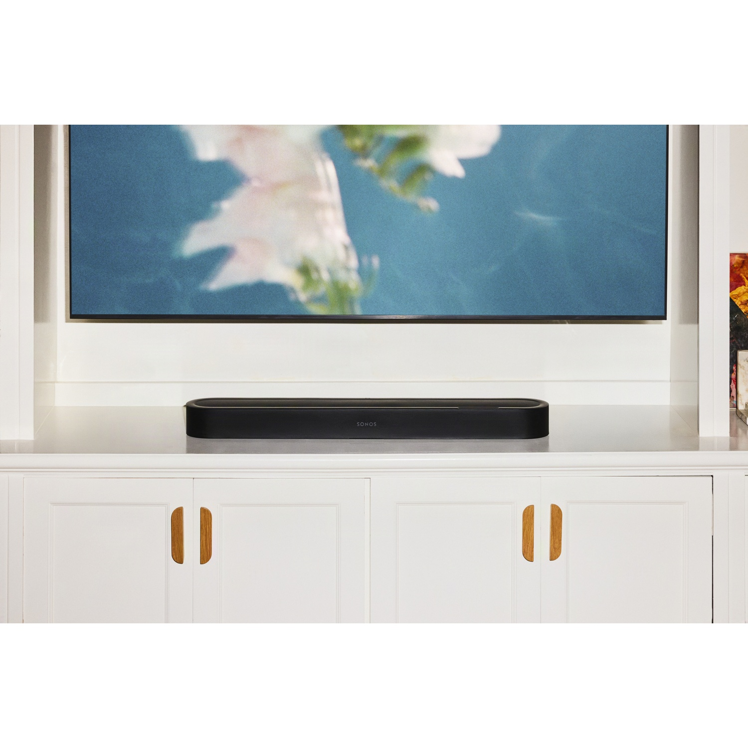 Une barre de son Sonos noire sur un meuble blanc sous une télévision affichant une image bleue.