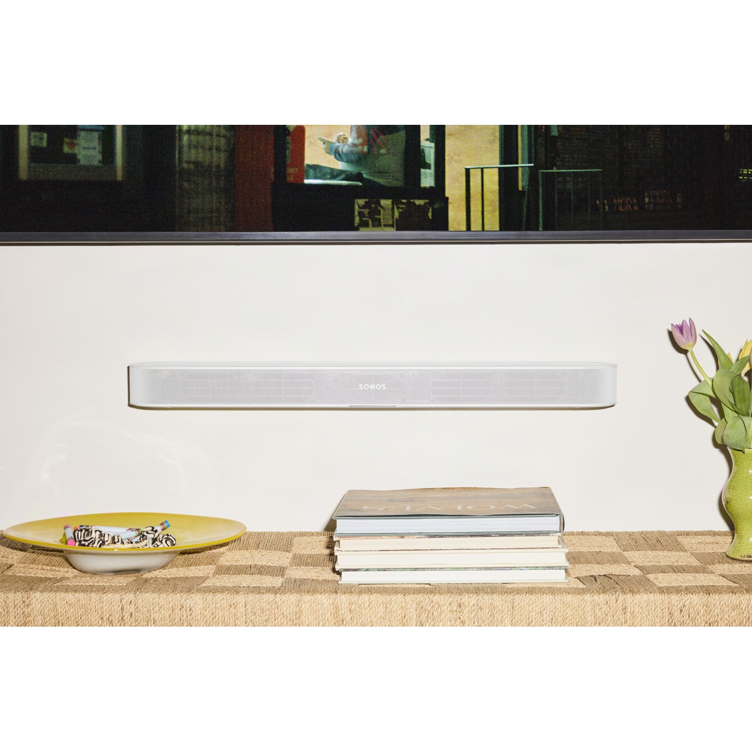 Une barre de son Sonos blanche est fixée au mur sous une télévision, avec des livres et un vase sur une table.