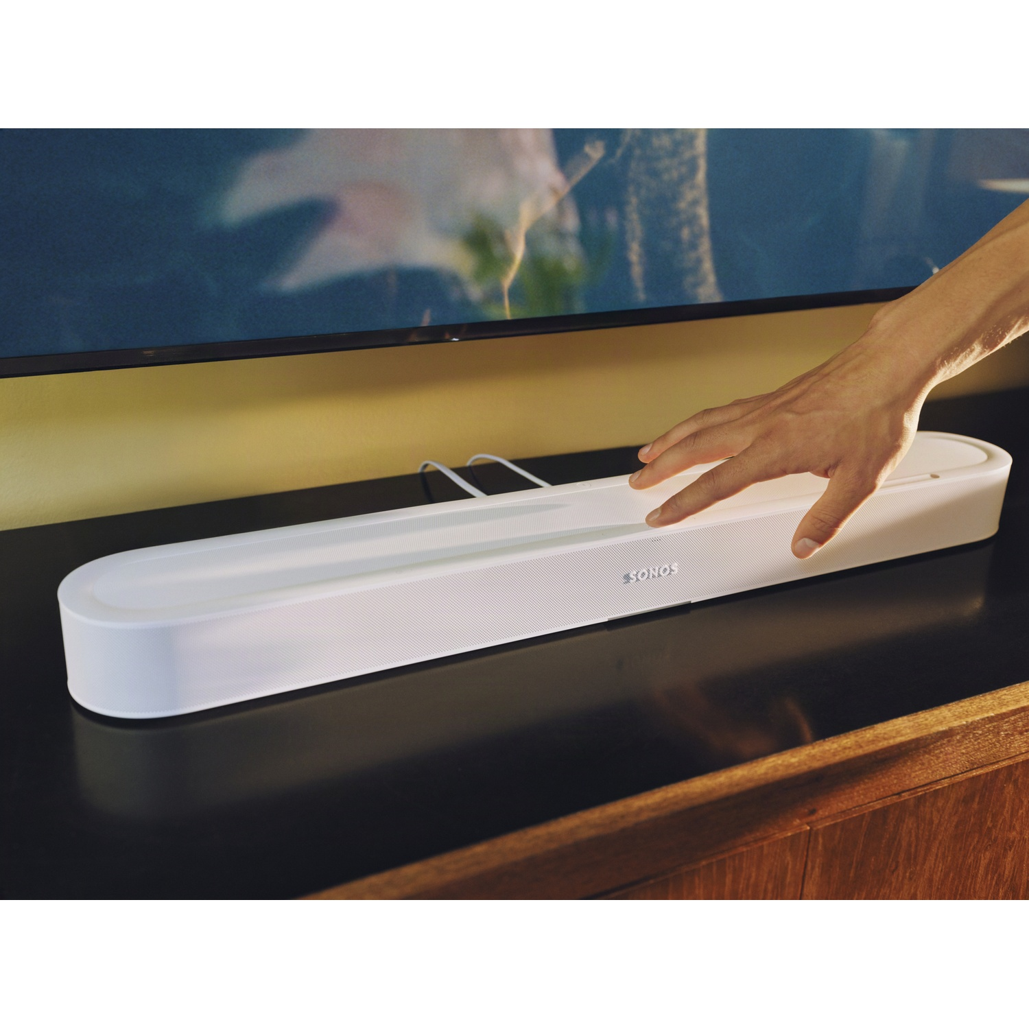 Une main touche une barre de son Sonos blanche posée sur une table sombre sous une télévision.