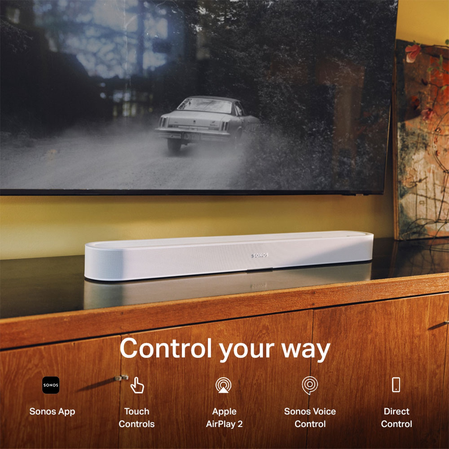 Une barre de son Sonos blanche repose sur un meuble en bois devant un téléviseur avec une voiture.