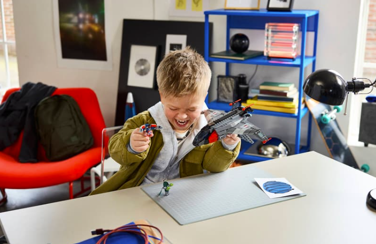 Ein junger Junge sitzt an einem Schreibtisch und spielt fröhlich mit LEGO-Figuren und einem Spielzeug-Raumschiff.