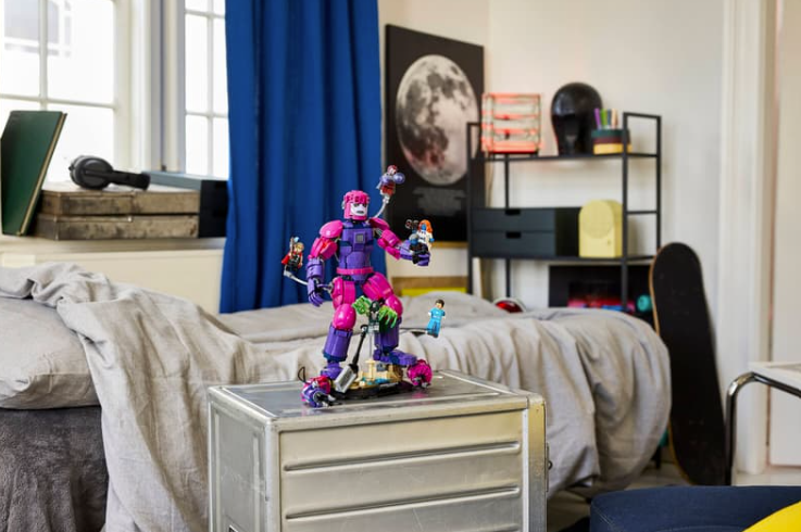 Eine pink-violette Lego-Roboterfigur steht auf einer Metallkiste in einem hellen Schlafzimmer.