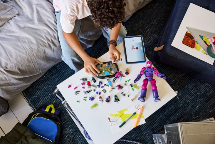 Ein Kind baut Lego-Figuren auf einem weißen Tisch neben einem Tablet.