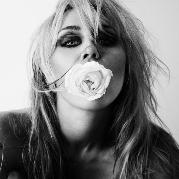 Schwarz-Weiß-Porträt einer blonden Frau mit dunklem Augen-Make-up, die eine weiße Rose im Mund hält.