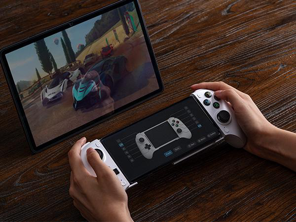 Jemand hält ein weißes Gamepad verbunden mit einem Smartphone auf einem Holztisch, daneben ein Tablet mit Rennspiel.