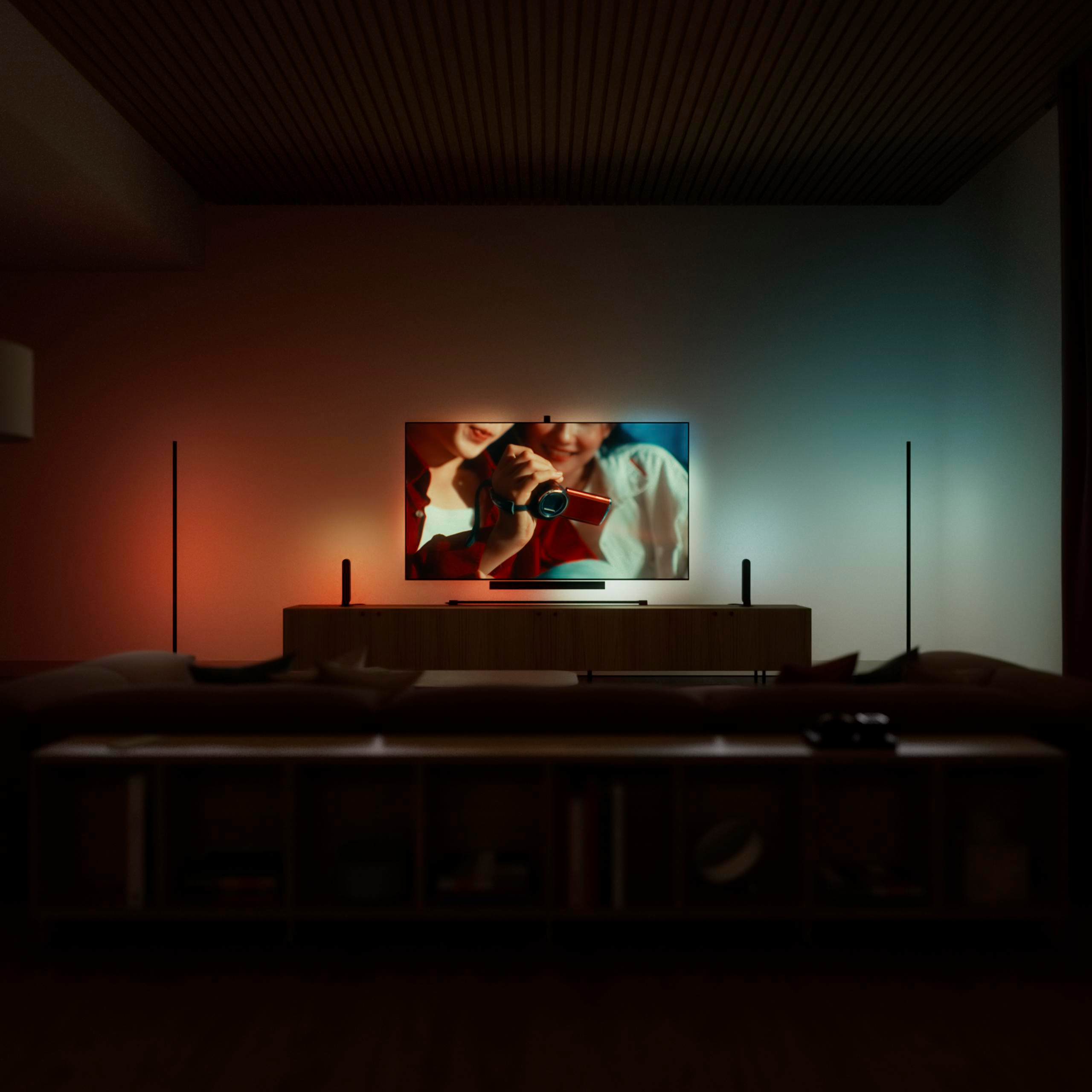 Wohnzimmer mit beleuchtetem Fernseher auf einem Schrank, flankiert von schlanken Lampen, und Regale im Vordergrund.