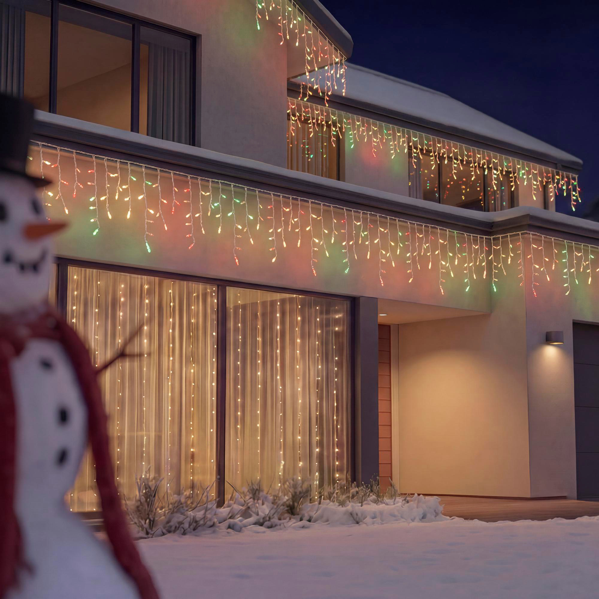 Ein verschneites modernes Haus nachts, geschmückt mit bunten Eiszapfenlichtern und einem Schneemann im Vordergrund.