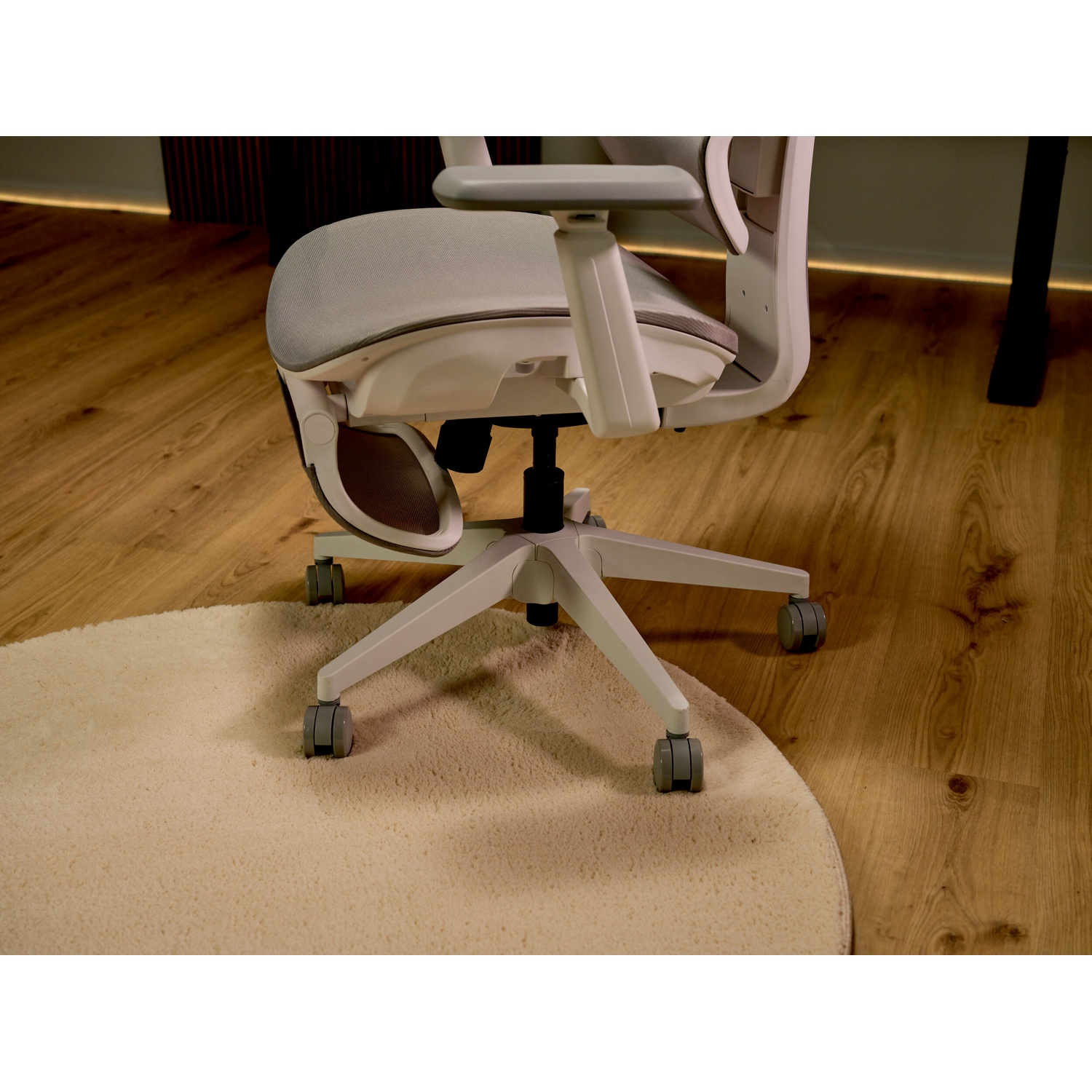 Una sedia da ufficchio ergonomica bianca con ruote si trova su un pavimento in legno con un tappeto rotondo chiaro.