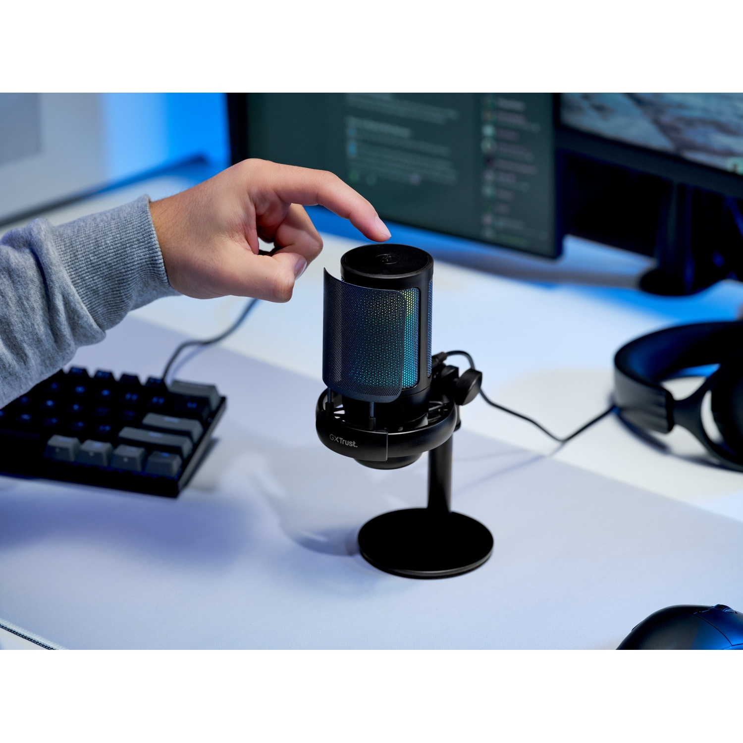 Una mano tocca un microfono nero con illuminazione RGB su una scrivania davanti ai monitor.