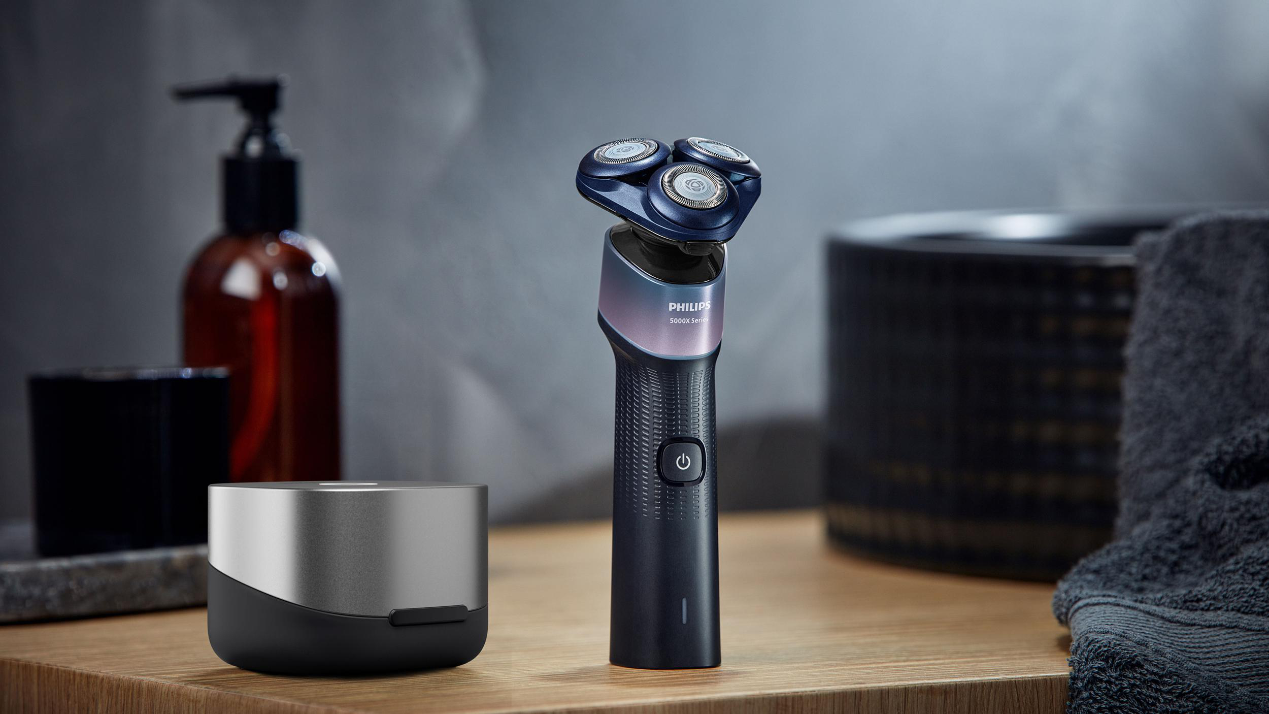 Philips 5000X Rasierer steht auf einer Holzoberfläche mit Ladeschale, Flasche und Handtuch im Hintergrund.