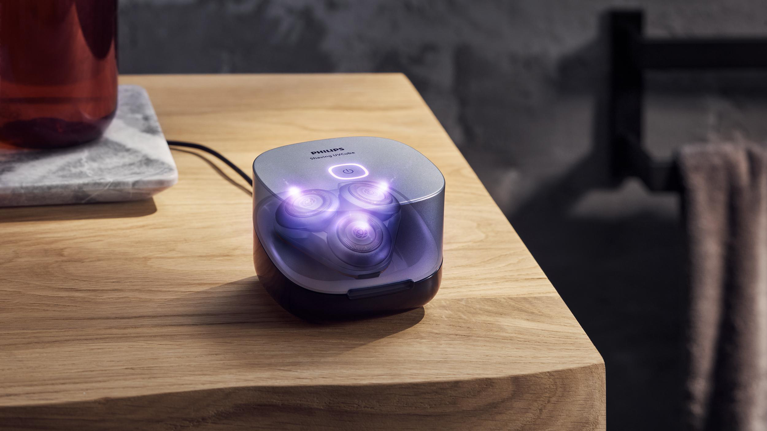 Ein Philips Shaving UVCube auf einem Holztisch, innen mit violettem UV-Licht beleuchtet, neben einem Marmoruntersetzer.
