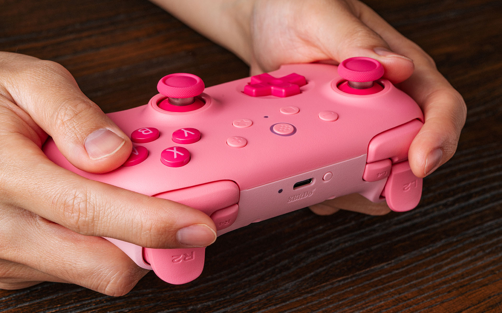 Hände halten einen rosa kabellosen Controller auf dunklem Holz.