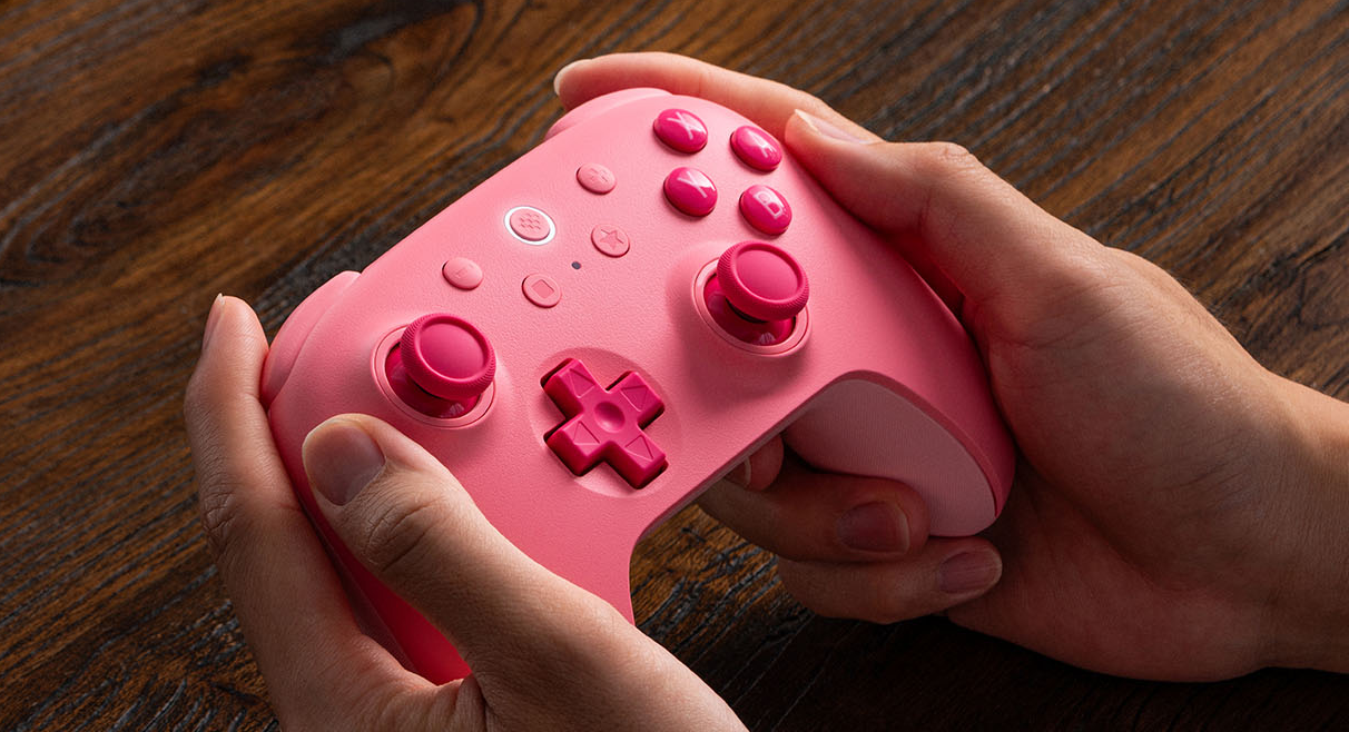 Hände halten einen pinken Videospiel-Controller auf einem hölzernen Hintergrund.