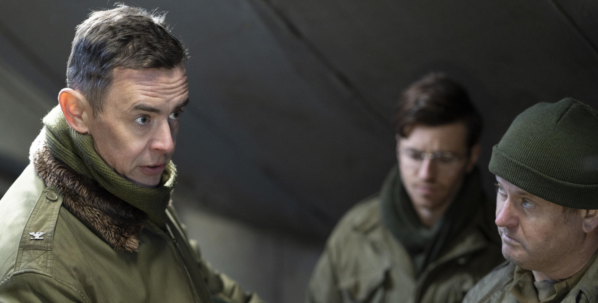 Drei Männer in olivgrünen Militäruniformen stehen in einem dunklen Raum zusammen.