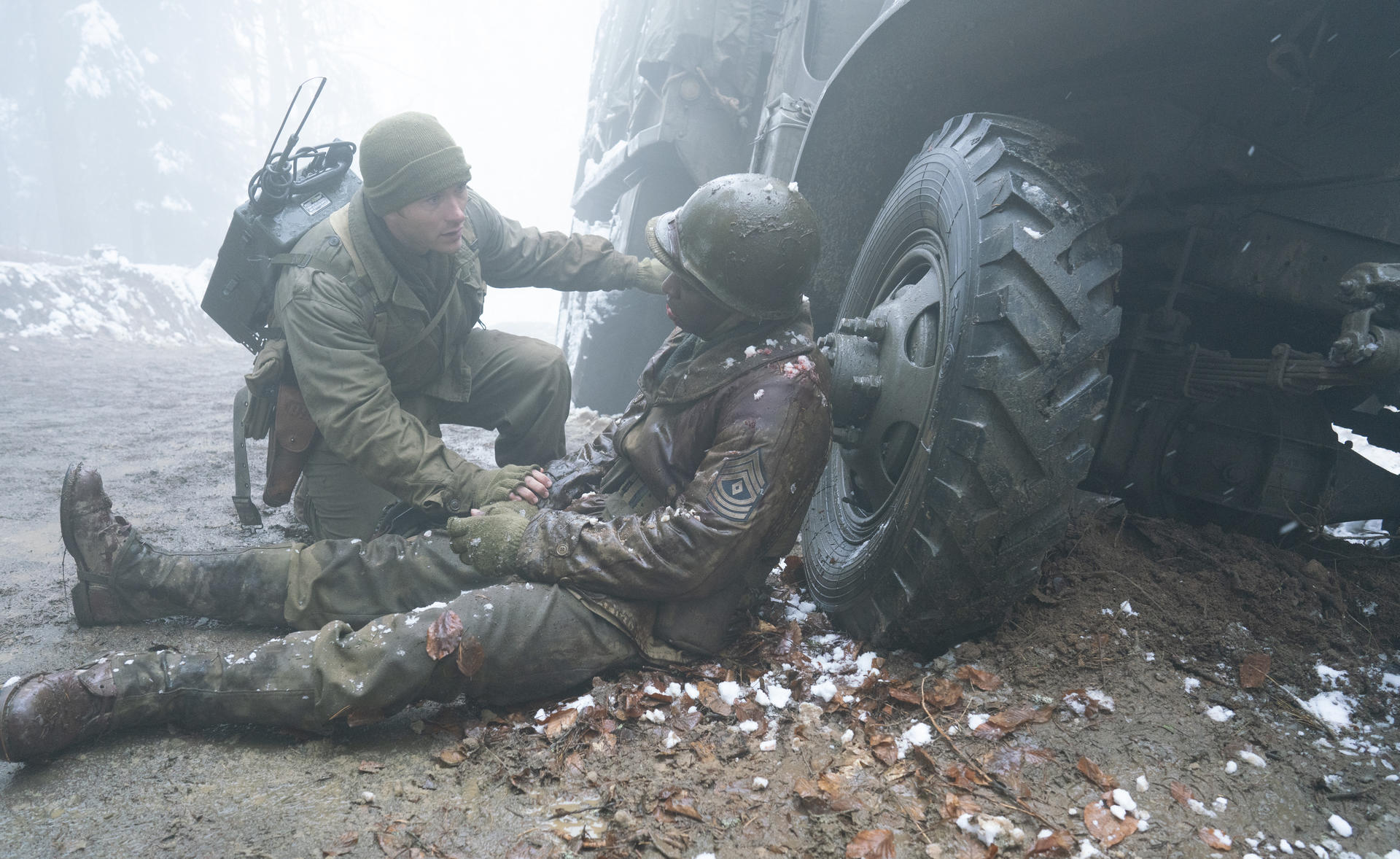 Ein Soldat in Winterkleidung hilft einem verletzten Soldaten im Schnee neben einem Lkw-Rad.