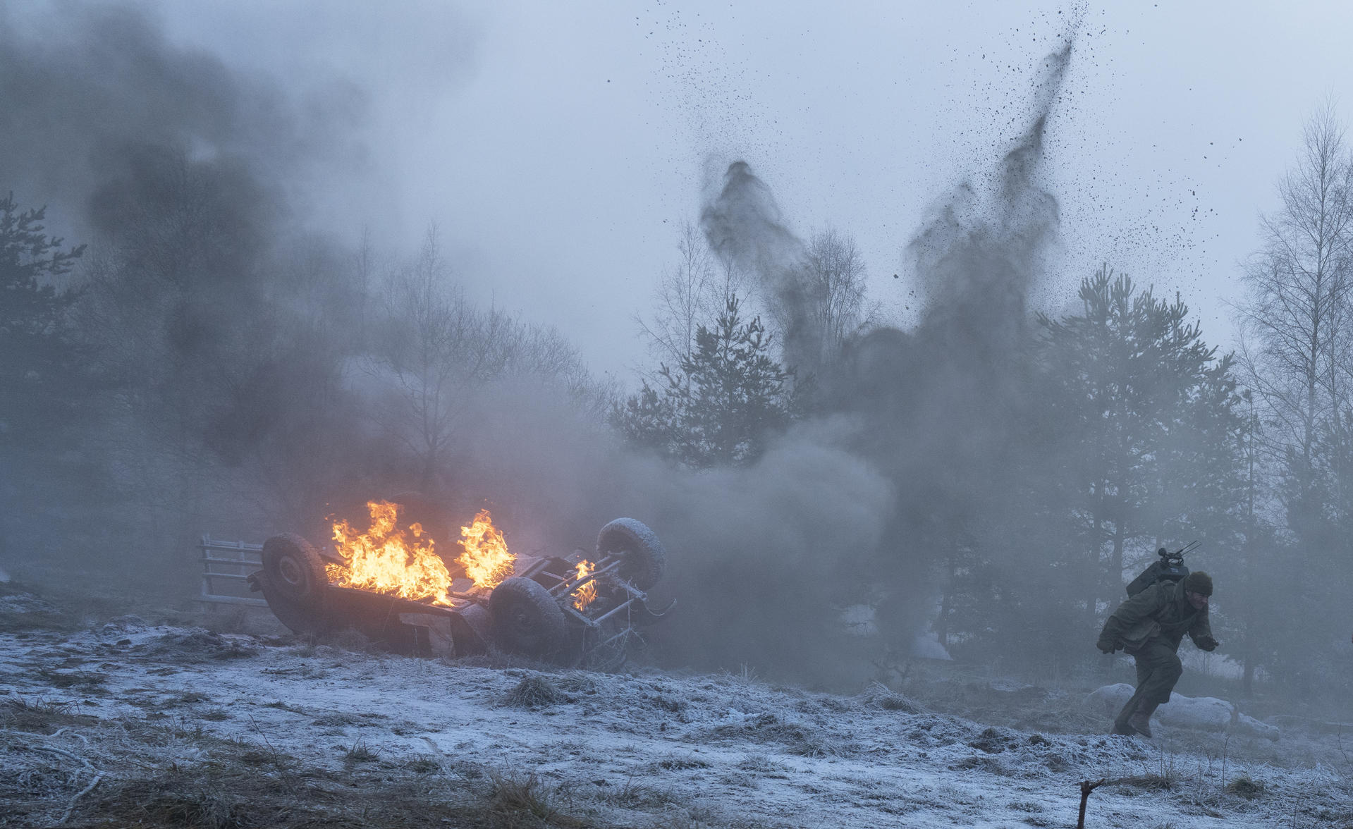 Ein brennendes, umgekipptes Auto in einer verschneiten Landschaft mit Rauch und einer Person, die wegläuft.