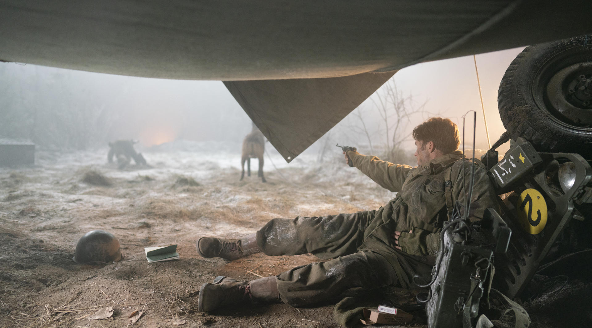 Ein Soldat in Uniform liegt unter einer Zeltplane im Schnee und zielt mit einer Pistole, neben ihm ein umgestürzter Jeep.