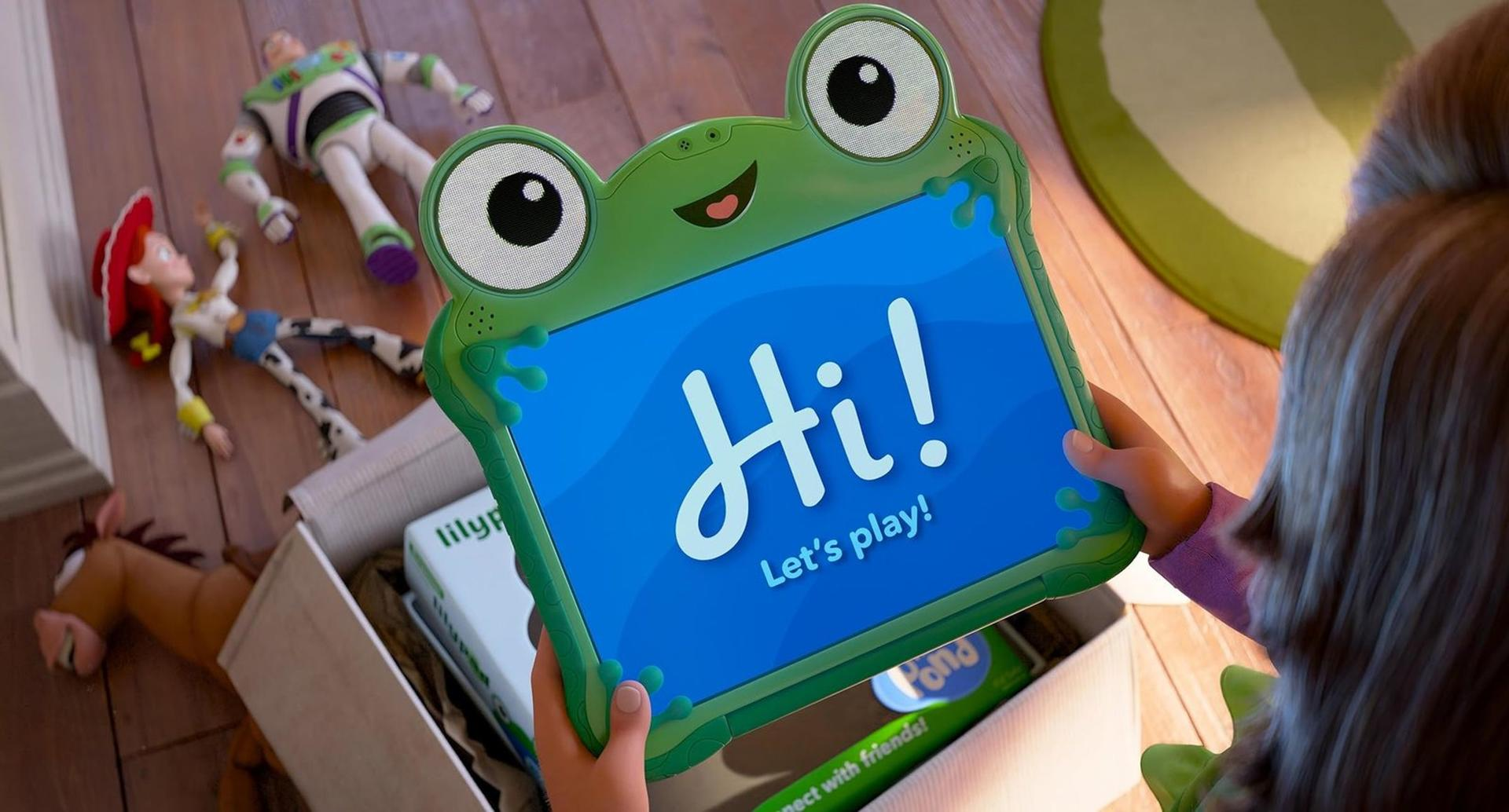 Ein Kind hält ein grünes Frosch-Tablet mit der Aufschrift 'Hi! Let's play!' in Händen, im Hintergrund liegen Spielsachen.