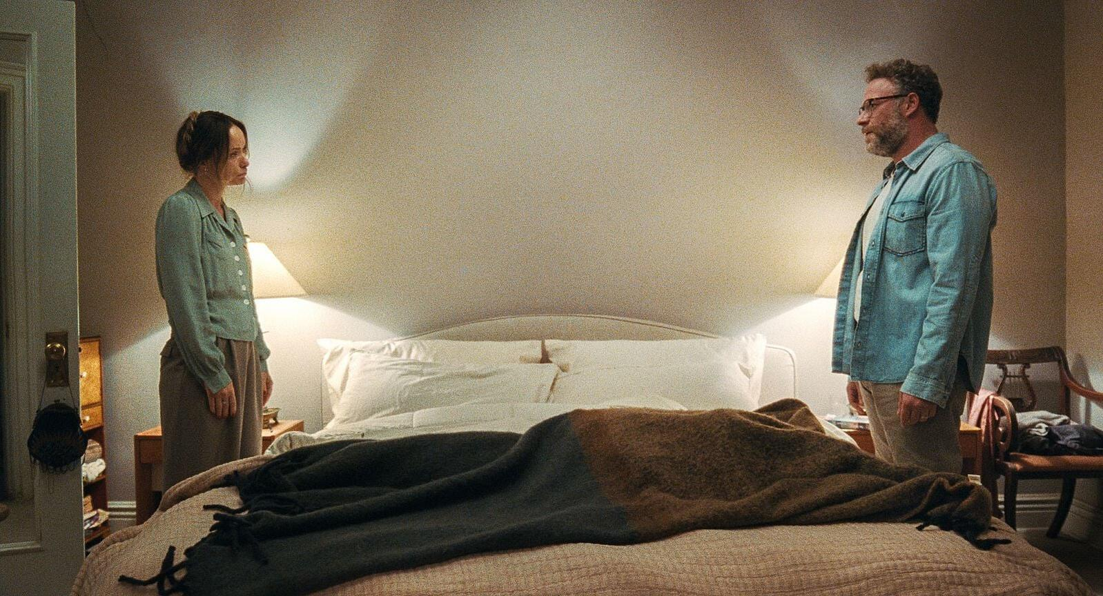 Ein Mann und eine Frau stehen sich in einem Schlafzimmer an den Seiten eines Bettes gegenueber.